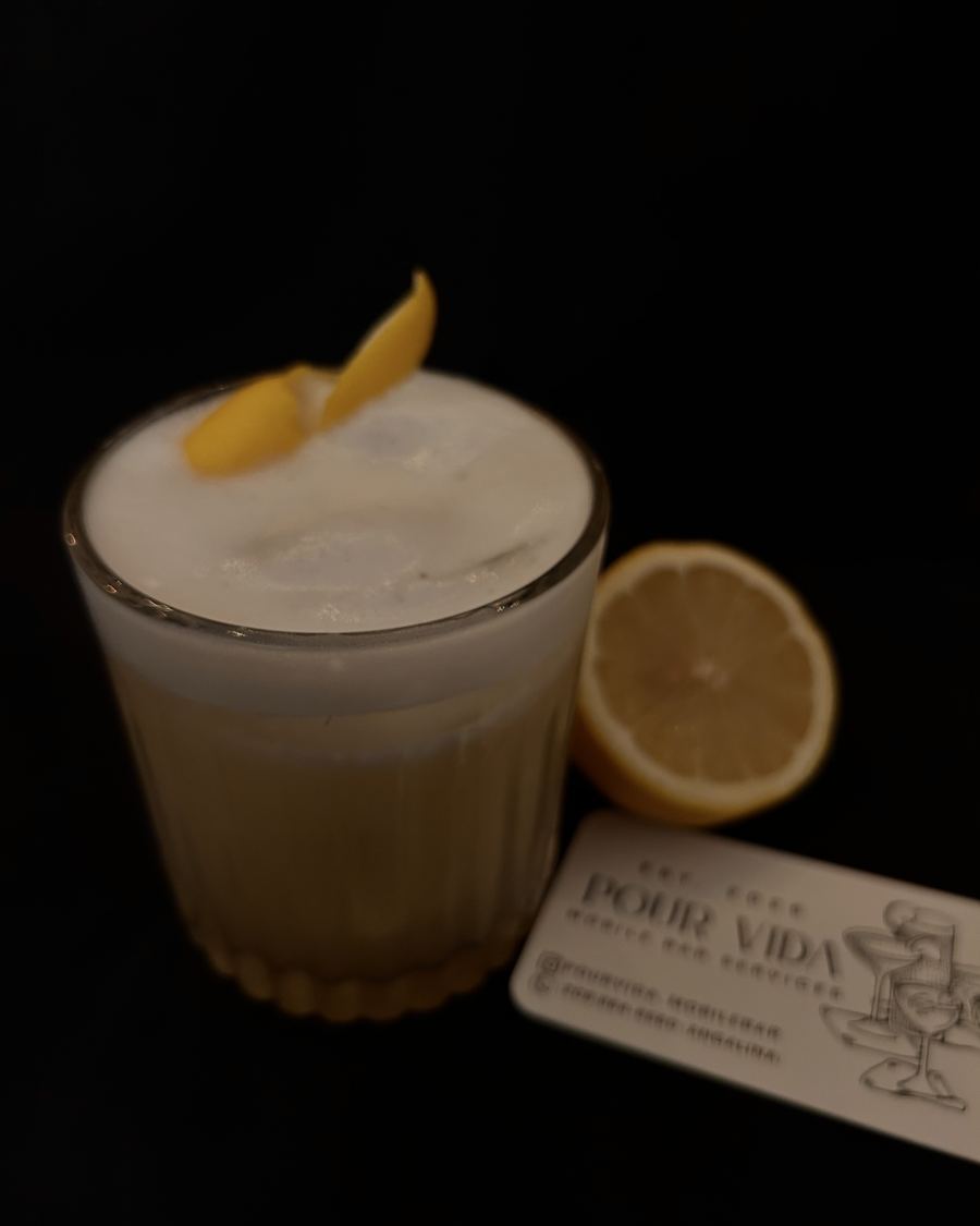 Whisky sour with a lemon peel garnish beside a lemon half and a Pour Vida card
